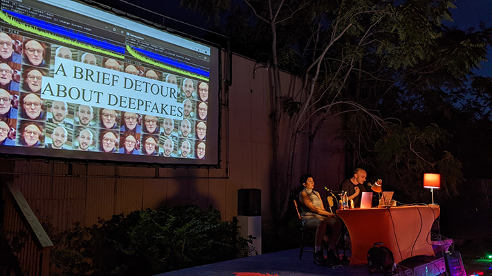 An outdoor stage with two presenters at a table infront of a projection screen with tiled images of morphed phases and the text 