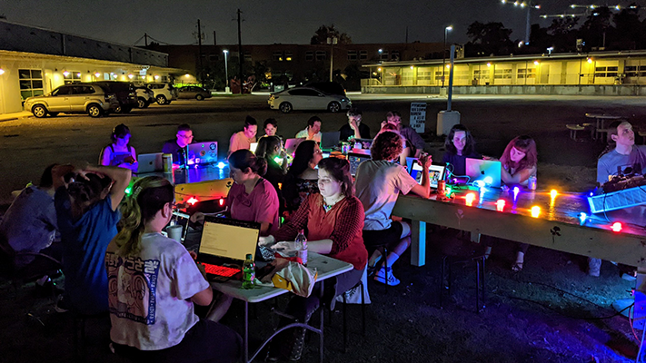 About 20 people seated around a large table with laptops, outdoors at night time. The table is illuminated with multi-color string lights.